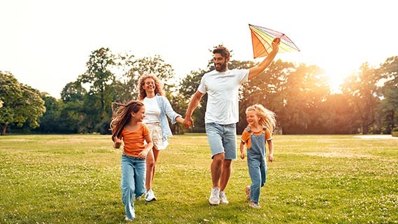 Happy young family dad, mom and two daughters running and playing with a colorful kite in a meadow in the park, having fun on a warm sunny weekend.