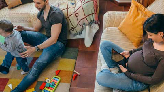 pregnant woman sitting on the couch looking at iPad next to partner playing with their toddler