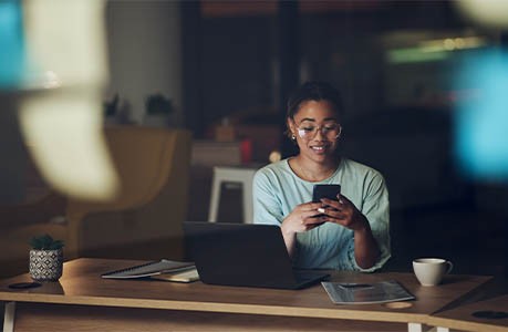 a woman sitting at a table using her laptop and phone in the dark