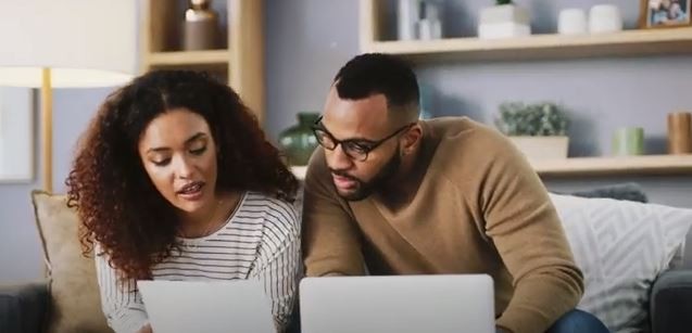 a man and woman sitting on the couch looking at their laptop